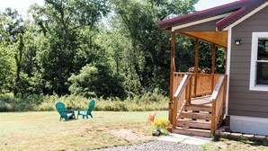 Une terrasse en bois avec un porche couvert, un chemin de gravier menant à un espace détente équipé de deux chaises, et une maison dont l’extérieur est recouvert de bardage brun.