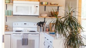 Une cuisine avec un four, un micro-ondes et des placards blancs. Il y a une étagère en bois au-dessus du micro-ondes et une plante en pot sur le sol.