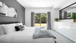 A modern bedroom with a large bed, grey curtains, a white dresser, and a view of greenery outside. - Marine Adventures – tranquil blue waters and white beaches of Jervis Bay (Callala Bay)