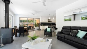A modern living room with a black leather sofa, a white coffee table, and a fireplace. There is a dining area with a table and chairs, and a kitchen in the background. - Marine Adventures – tranquil blue waters and white beaches of Jervis Bay (Callala Bay)