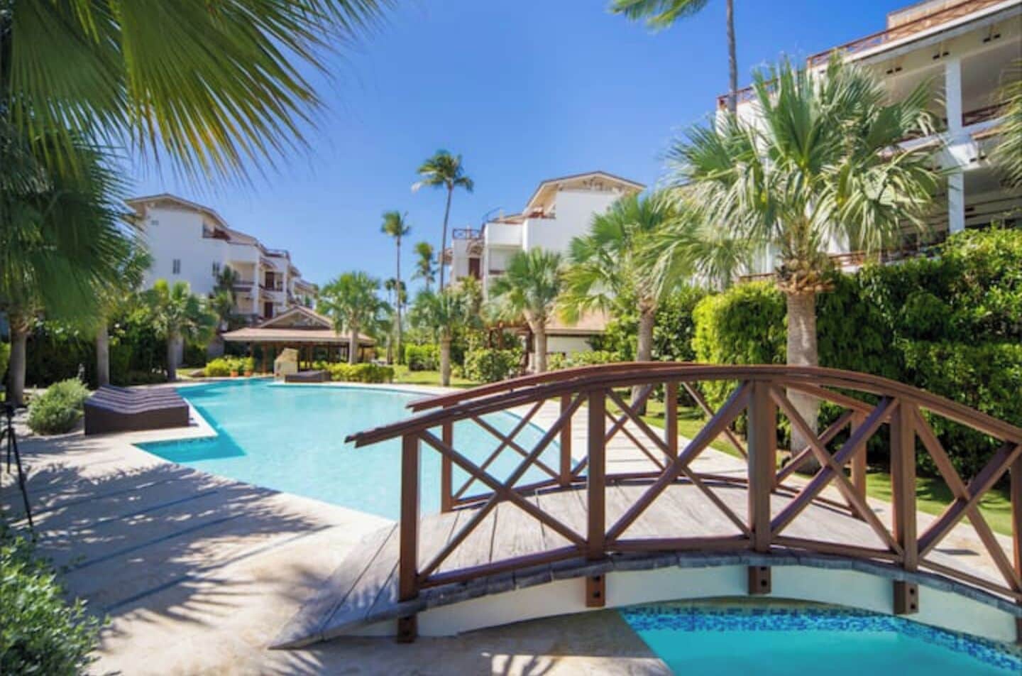 A resort with a large swimming pool, palm trees, and a wooden bridge.