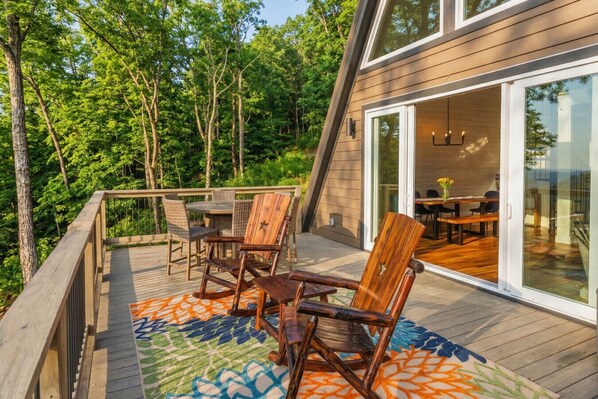 A wooden deck with outdoor furniture, a glass door leading to a dining area, and a forested background. - A-Frame Cabin (Independence) - by Red White and Blue Views (Lookout Mountain)