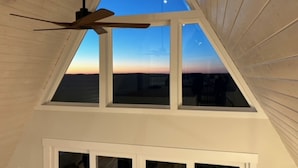 Et værelse med skrå væg, loftventilator og udsigt til solnedgang gennem et ovenlysvindue.