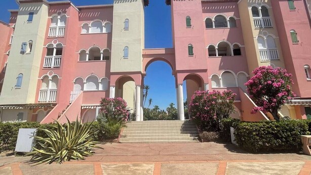 A multi-story hotel with a central archway, pink and beige exterior, and steps leading to the entrance. - Superb modern 2/4-person apartment with swimming pool (Le Barcarès)
