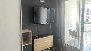 A modern living room with a flat-screen TV mounted on the wall, a wooden cabinet with drawers, a small table with chairs, and a sliding glass door leading to an outdoor area. - Superb modern 2/4-person apartment with swimming pool (Le Barcarès)