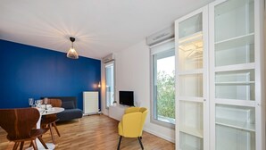 Una sala moderna con pared azul de acento, piso de madera, zona de comedor con mesa y sillas, un sillón amarillo, un televisor sobre un mueble blanco y un ventanal con vistas.