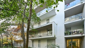 Un edificio de varios pisos con balcones, una entrada porticada y abundante vegetación.
