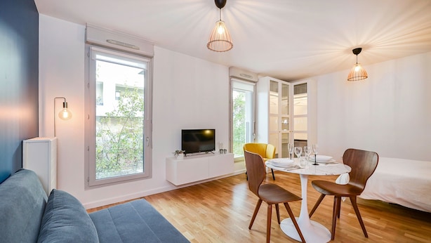 Una sala moderna con un sofá, una mesa de comedor con sillas, un televisor y ventanas grandes.