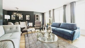 Ein modernes Wohnzimmer mit dunkler Akzentwand, einem blauen Sofa, einem grauen Sessel, einem runden Glastisch und einer Lampe.