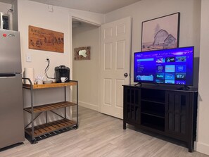 A kitchen with a refrigerator, coffee maker, and a cart. A TV is mounted on a cabinet displaying a streaming service interface. - Cozy studio walking distance to downtown Fort Myers (Fort Myers)