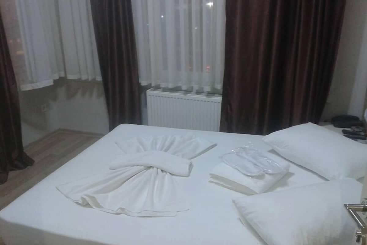 A neatly made bed with white linens and pillows, a bedside table with a lamp, and a window with curtains.