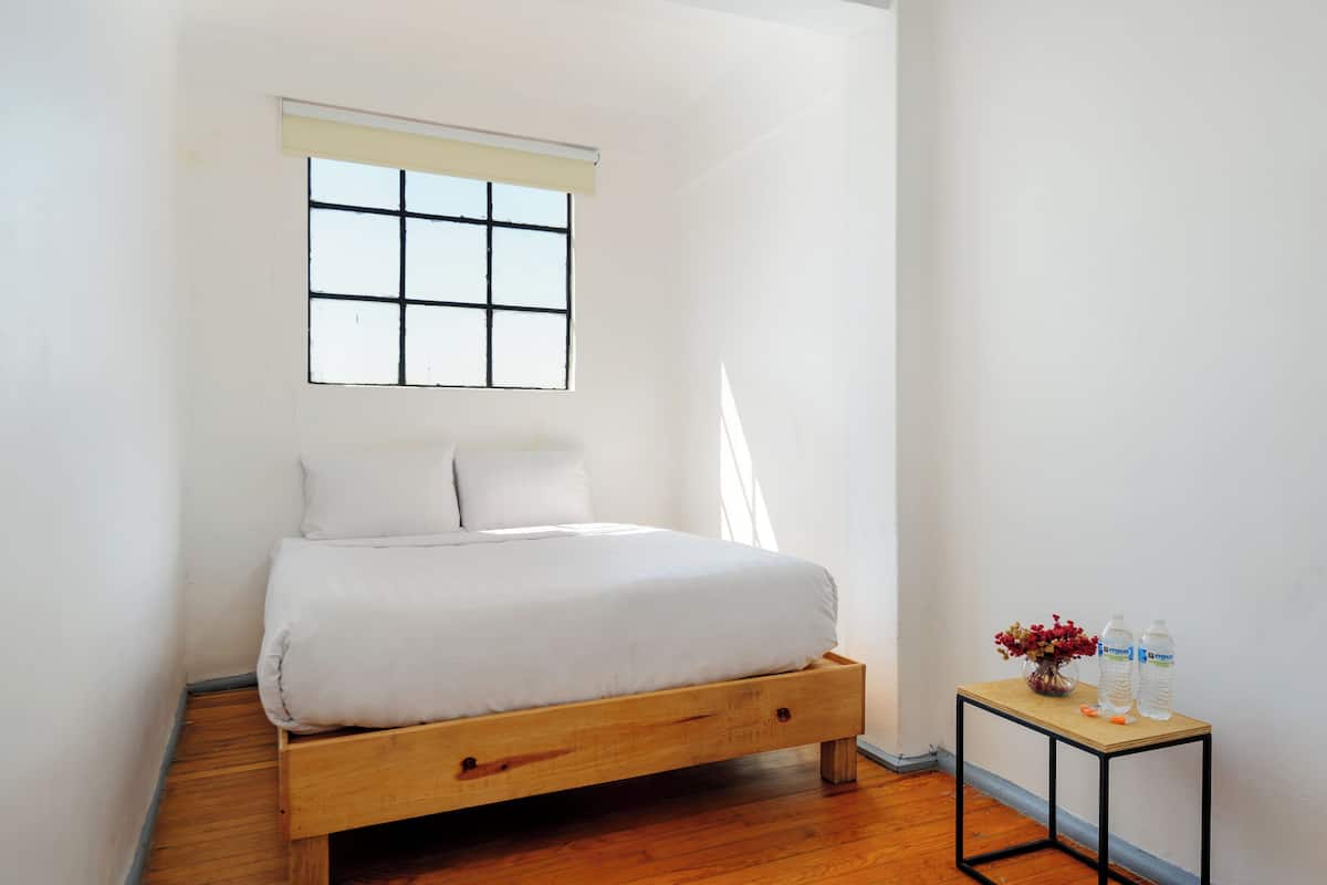 A minimalist bedroom with a wooden bed, a small table with water bottles, and a window with blinds.
