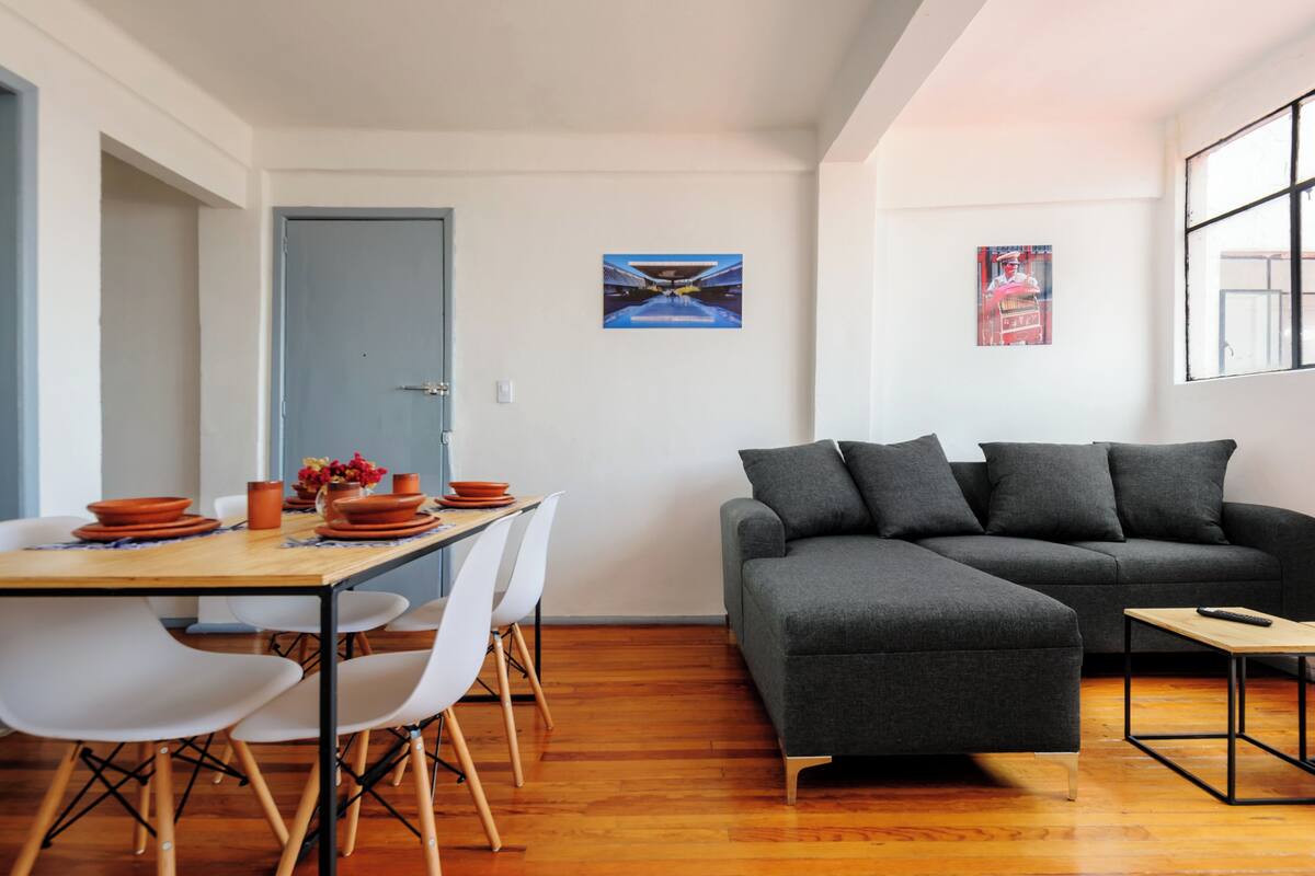 A modern living room with a grey sectional sofa, a wooden dining table with white chairs, and a small coffee table.