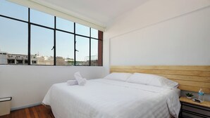 Una habitación de hotel con un ventanal grande, un cabecero de madera, una cama bien hecha, una mesita de noche con un control remoto y una planta pequeña.