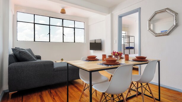 Una sala de estar moderna con sofá, mesa de comedor y sillas. Hay un ventanal grande, un televisor de pantalla plana y un espejo decorativo en la pared.