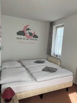 Quarto com duas camas, abajur vermelho, radiador e janela com cortinas.