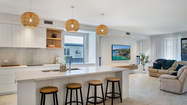 Una cocina moderna con una isla central, taburetes de bar, luces colgantes y un amplio ventanal.