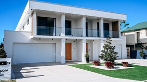 A modern two-story house with a white facade, a balcony on the upper floor, and a well-maintained lawn with red plants.