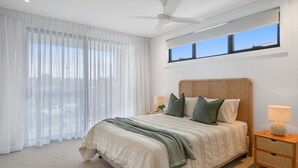 Un dormitorio con una cama grande, un ventilador de techo, una ventana con persianas y una mesita de noche con lámpara.