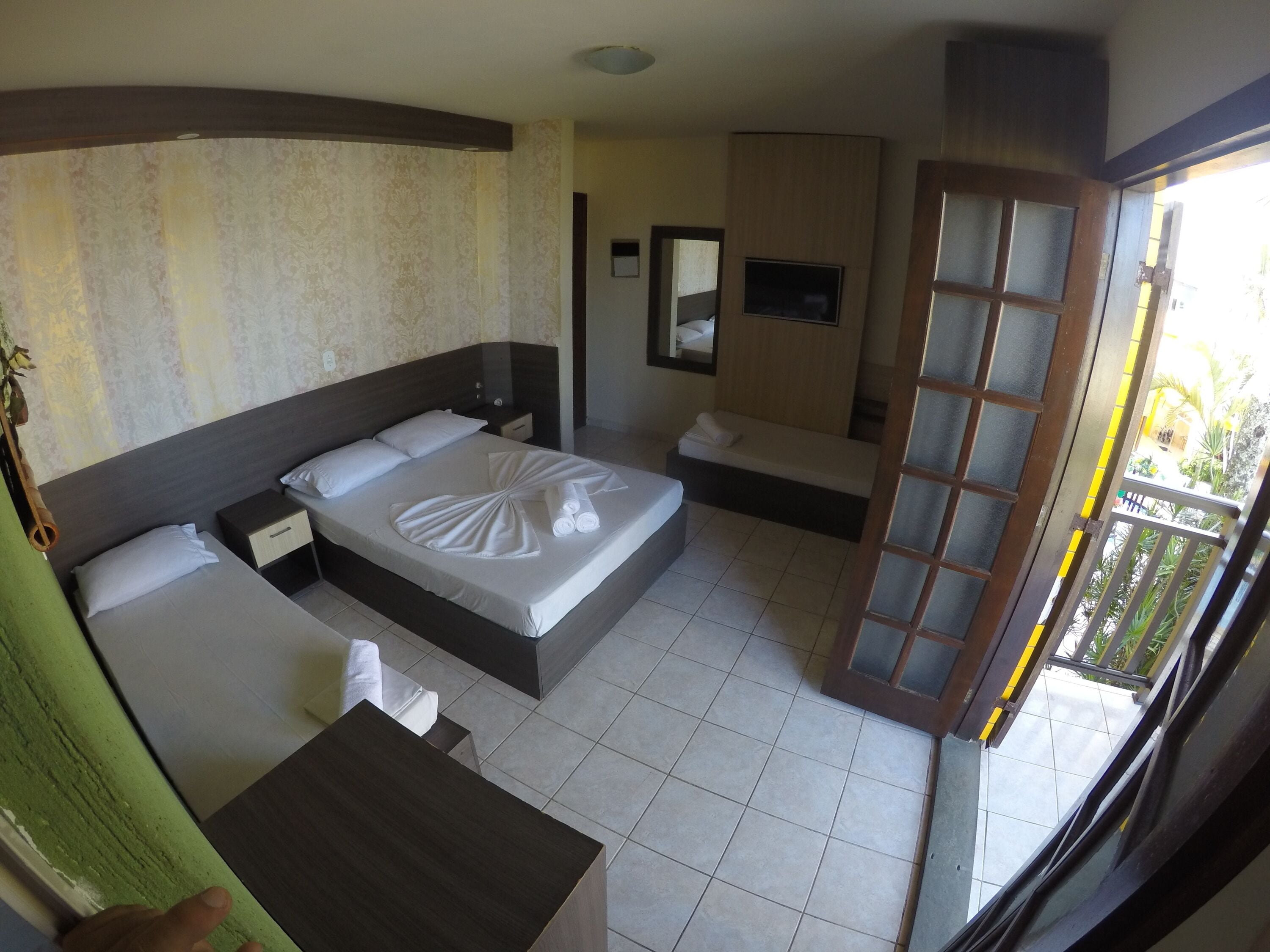 A hotel room with a bed, bedside tables, a small desk, a mirror, and a window with a view of greenery.