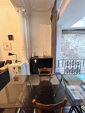 A room with a glass table, a wooden chair, a black sofa, a small kitchen area, and a view of a brick wall. - Chic Comfortable Loft and Natural Light (Brussels)