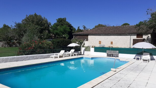 A rectangular swimming pool with a tiled edge, surrounded by a white tiled deck. There are white lounge chairs and umbrellas, and a stone building with a tiled roof in the background. - Gite du Fourbas - renovated in 2025 - with swimming pool (Belmont-Sainte-Foi)