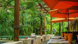 Een tropisch resort met een houten terras, ligbedden en een buitenzwembad, omringd door weelderige begroeiing.