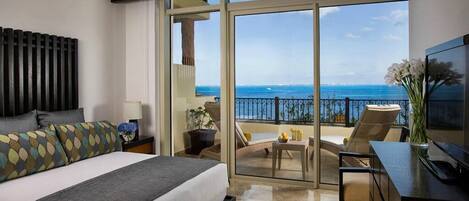 A hotel room with a large bed, a TV, a balcony with a view of the sea, and a dining area.