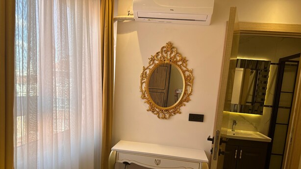Quarto de hotel com escrivaninha branca, espelho com moldura dourada e banheiro com pia e espelho.