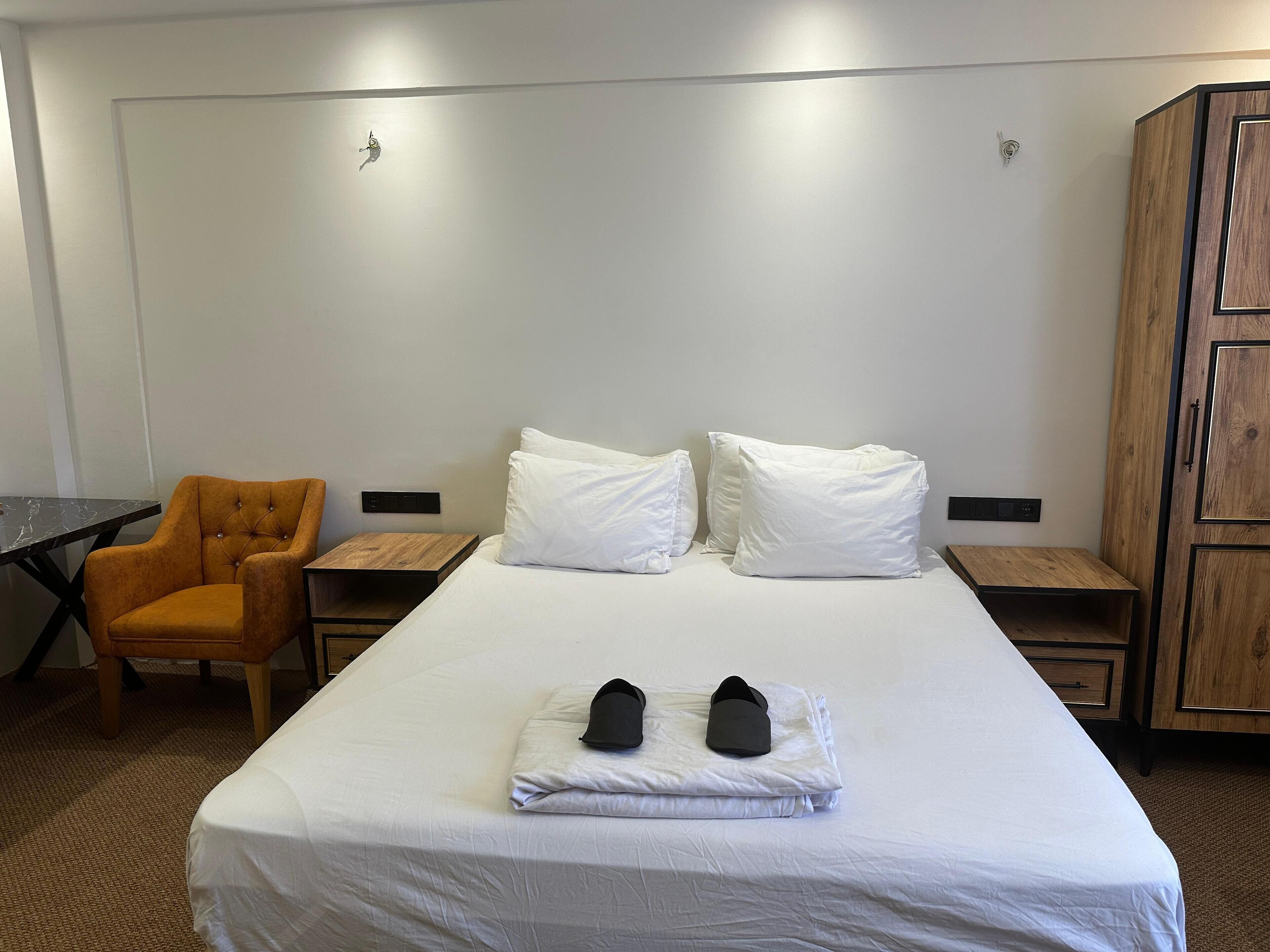 A hotel room with a large bed, two bedside tables, a wooden wardrobe, a small wooden nightstand, and an orange armchair.