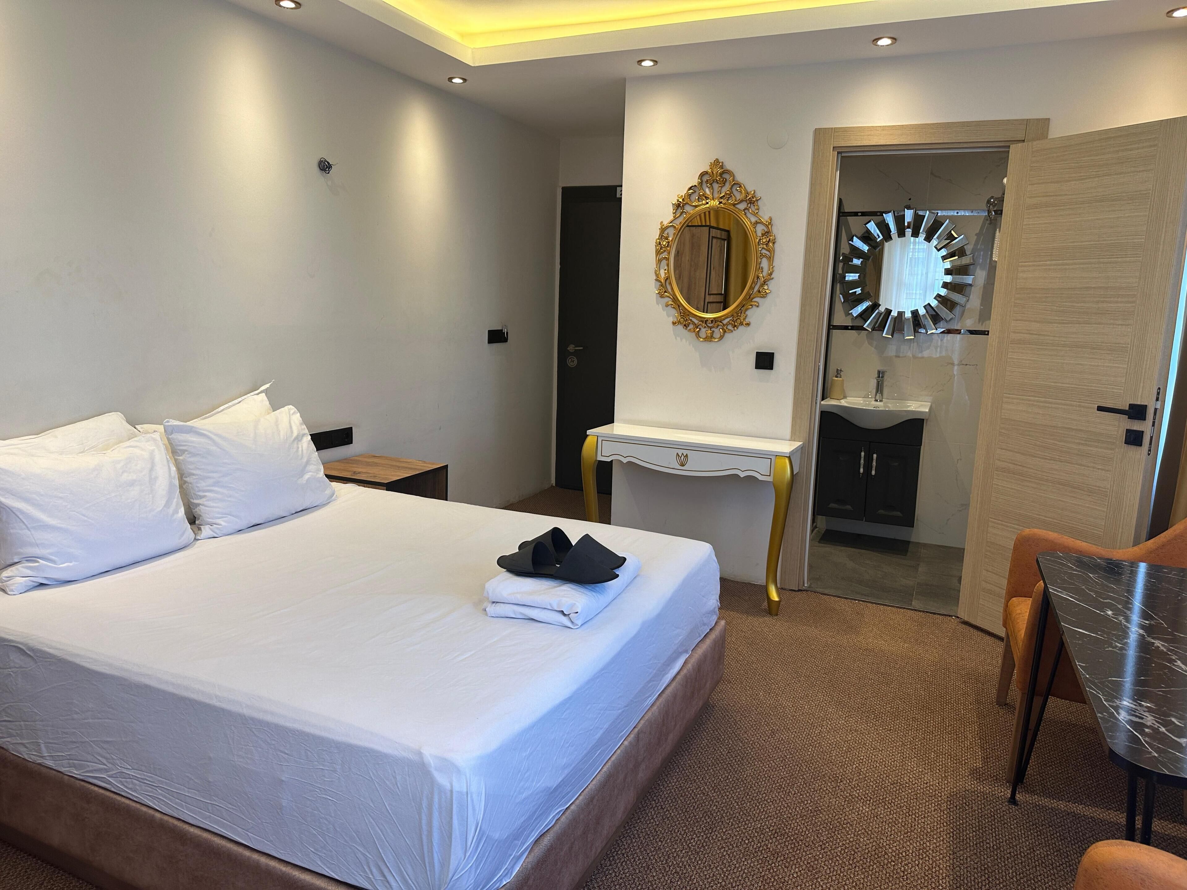 A hotel room with a large bed, a bedside table, a mirror, a vanity with a sink, and a door leading to another room.