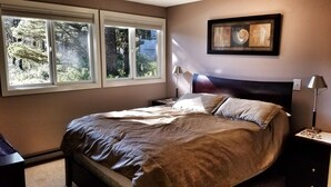 A bedroom with a bed, two bedside tables, a nightstand, a window with a view of trees, and a framed artwork on the wall.