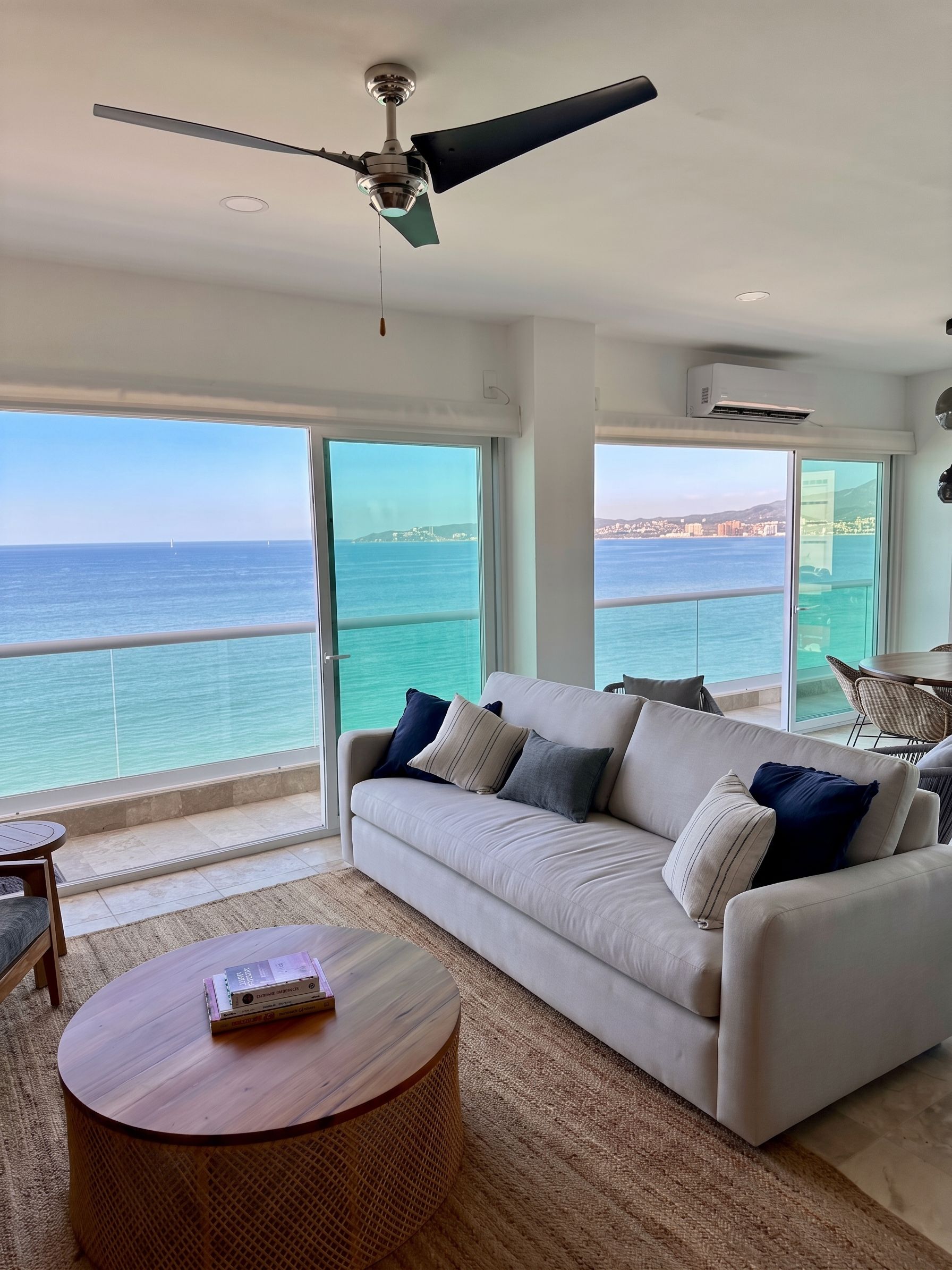 A modern living room with a large window overlooking the sea, a ceiling fan, and a round wooden table with books.