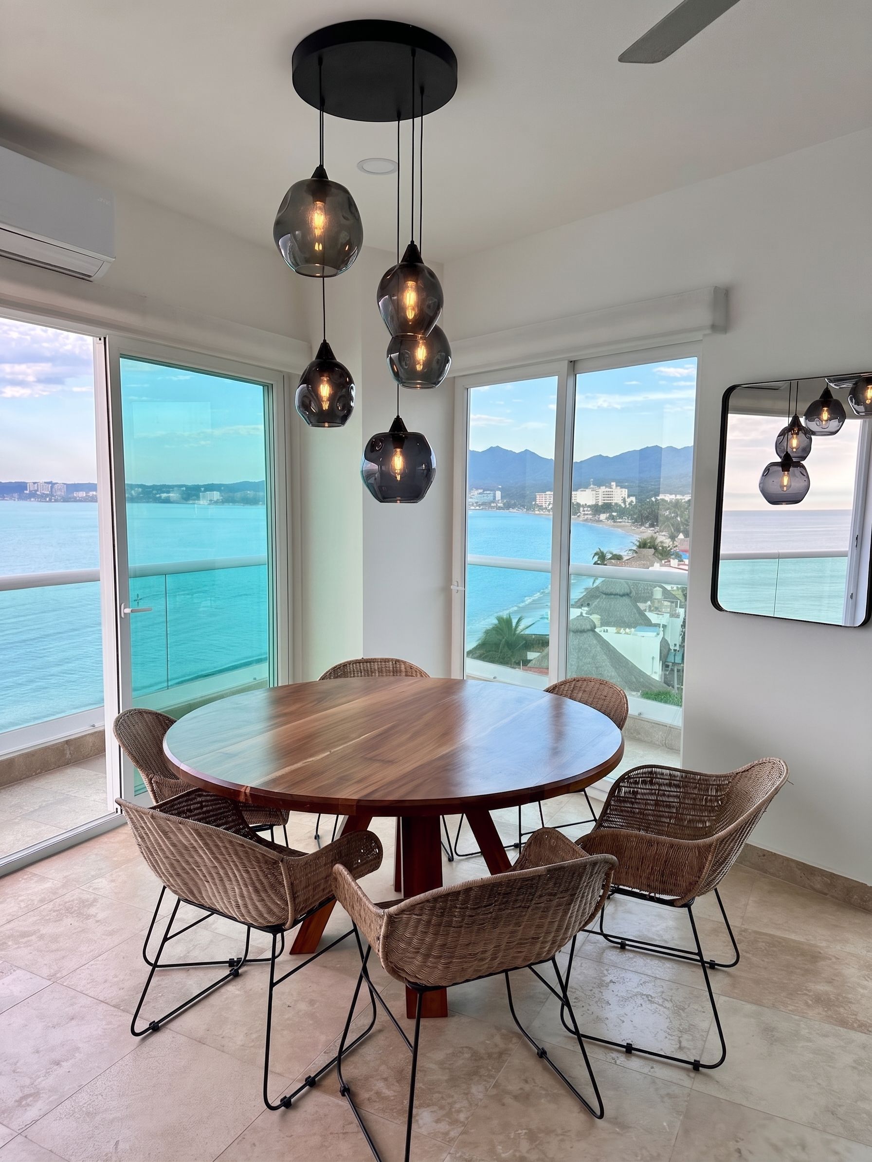 A round dining table with wicker chairs, a modern pendant light fixture, and a view of the sea through large windows.