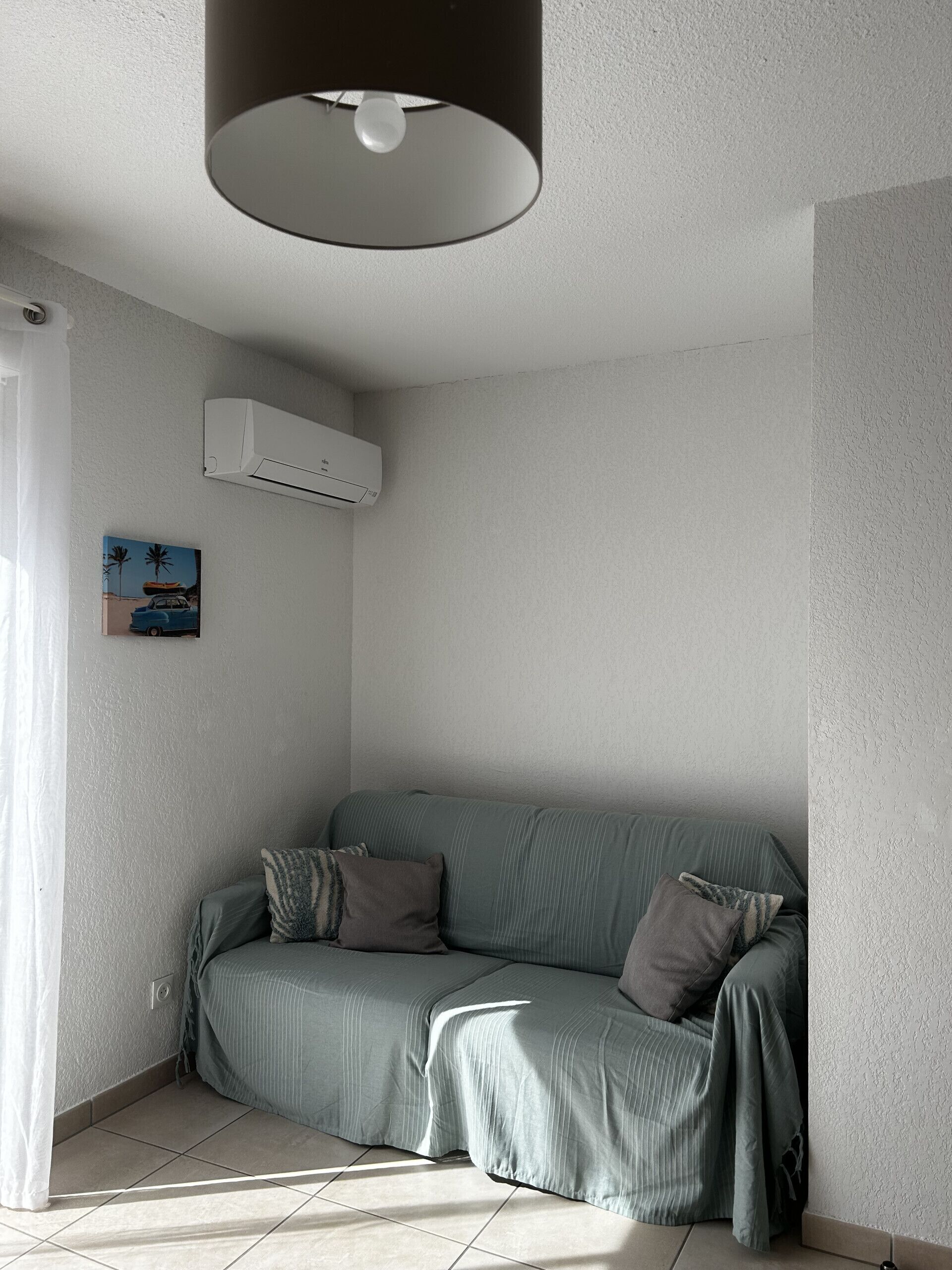 Quarto de canto com sofá cinzento, luminária no teto, ar-condicionado e um quadro na parede.