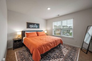Quarto com cama, mesas de cabeceira, tapete, arte com tema de motocicleta, janela com persianas e vista para casas lá fora.