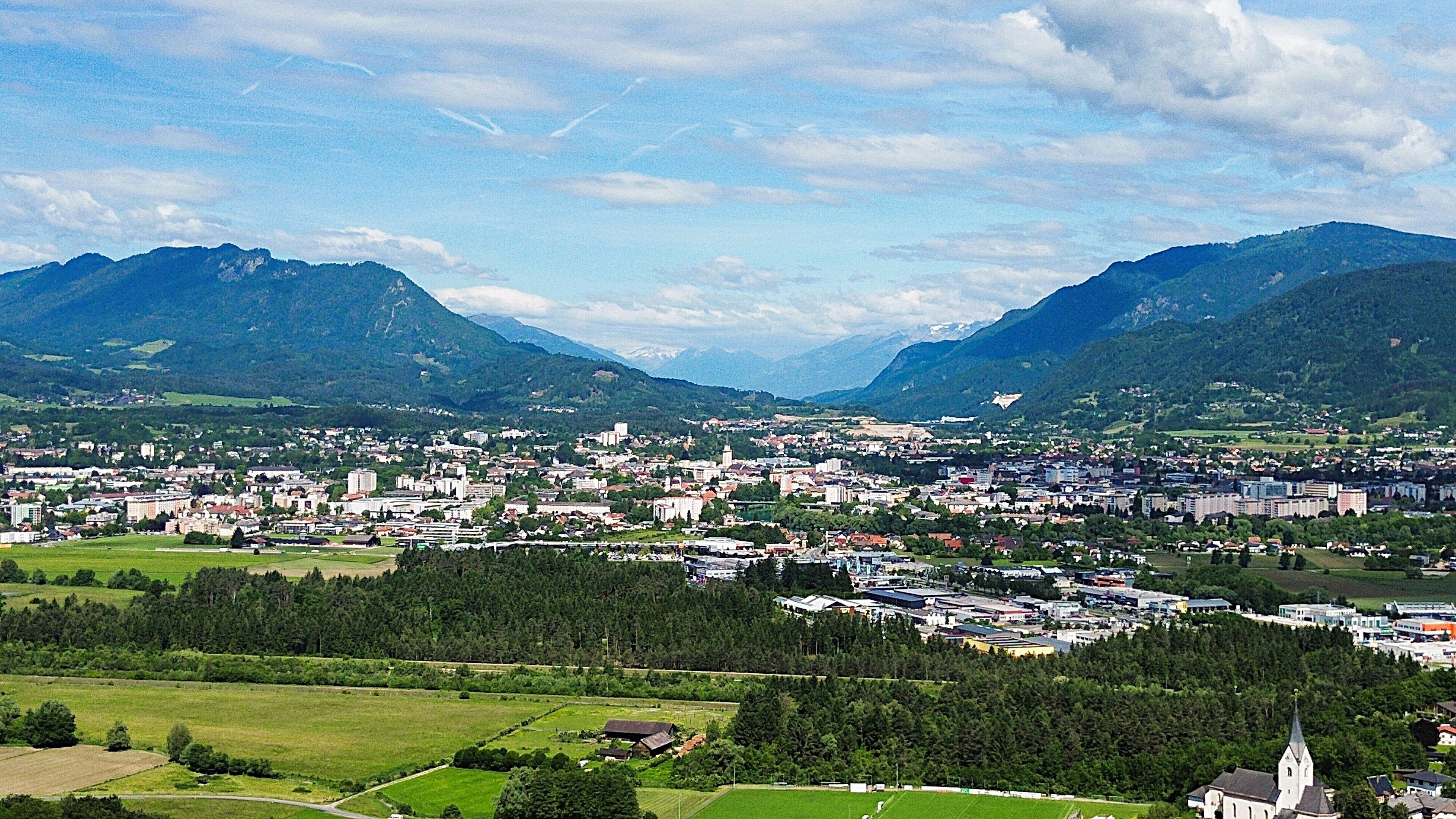 A town nestled between mountains with a mix of residential and commercial buildings, surrounded by green fields and forests.