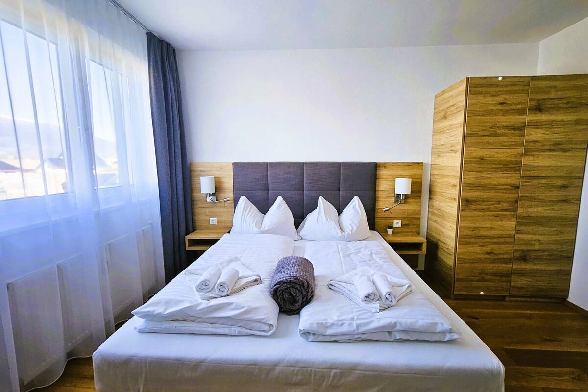 Une chambre d’hôtel avec un grand lit, deux tables de chevet avec des lampes, une armoire en bois et une fenêtre avec des rideaux légers.