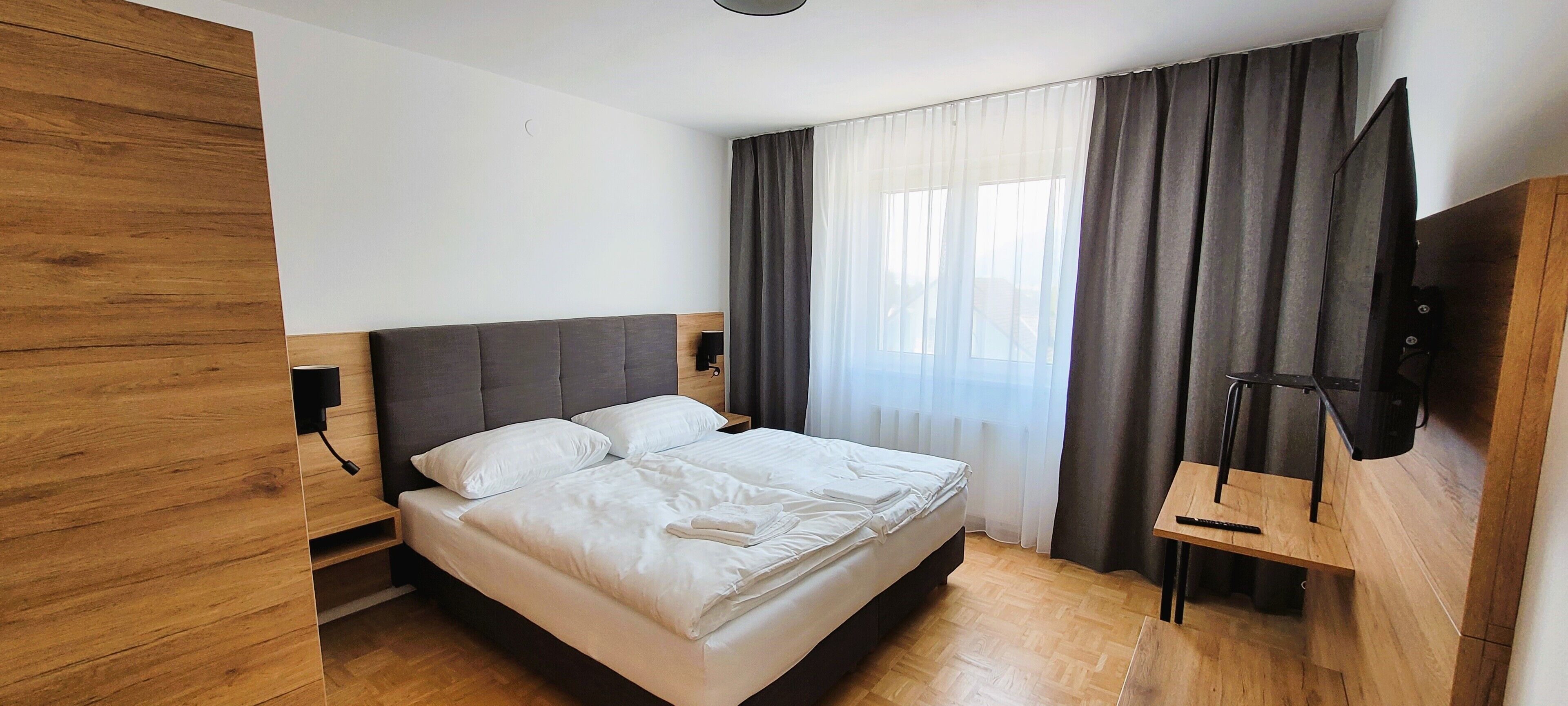 Une chambre à coucher avec un lit, une tête de lit en bois, un téléviseur fixé au mur et une fenêtre avec des rideaux.
