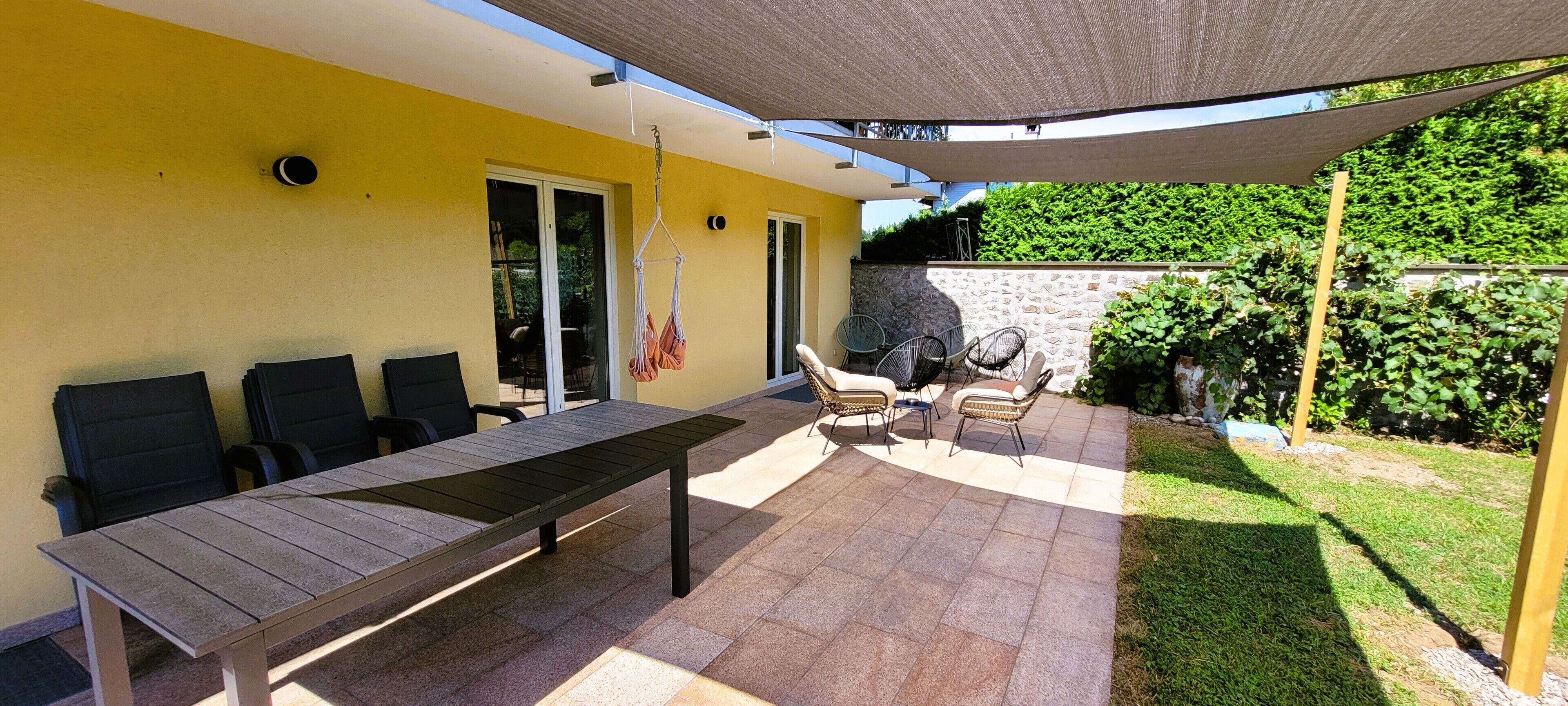 An outdoor patio with a long table and several chairs, a hanging chair, and a covered area with a canopy.