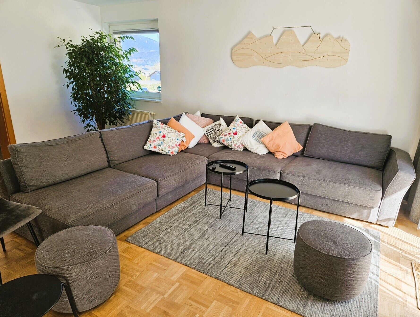Un salon moderne avec un canapé d’angle gris, deux tables d’appoint noires, un tapis gris et une grande fenêtre donnant sur une montagne.