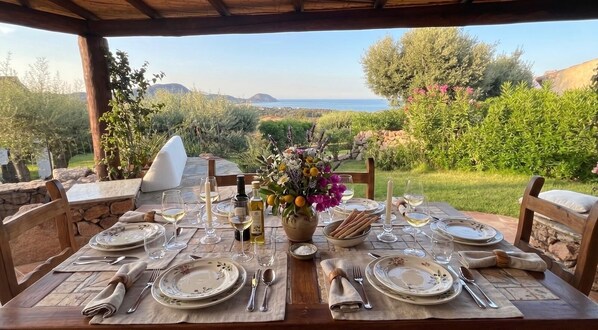 Un área de comedor al aire libre con una mesa preparada para cuatro personas, rodeada de vegetación y vistas al mar.