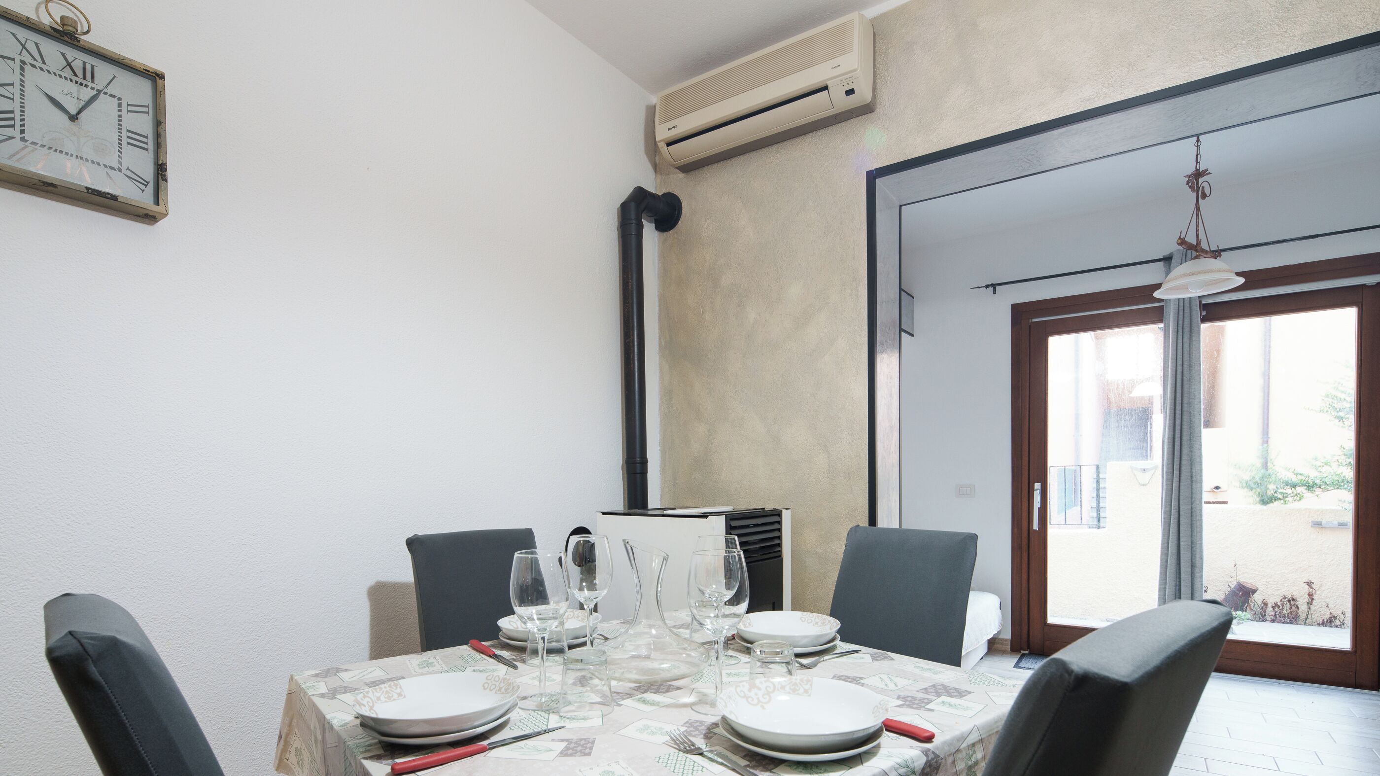 A dining area with a table set for four, a clock on the wall, and an air conditioning unit above.
