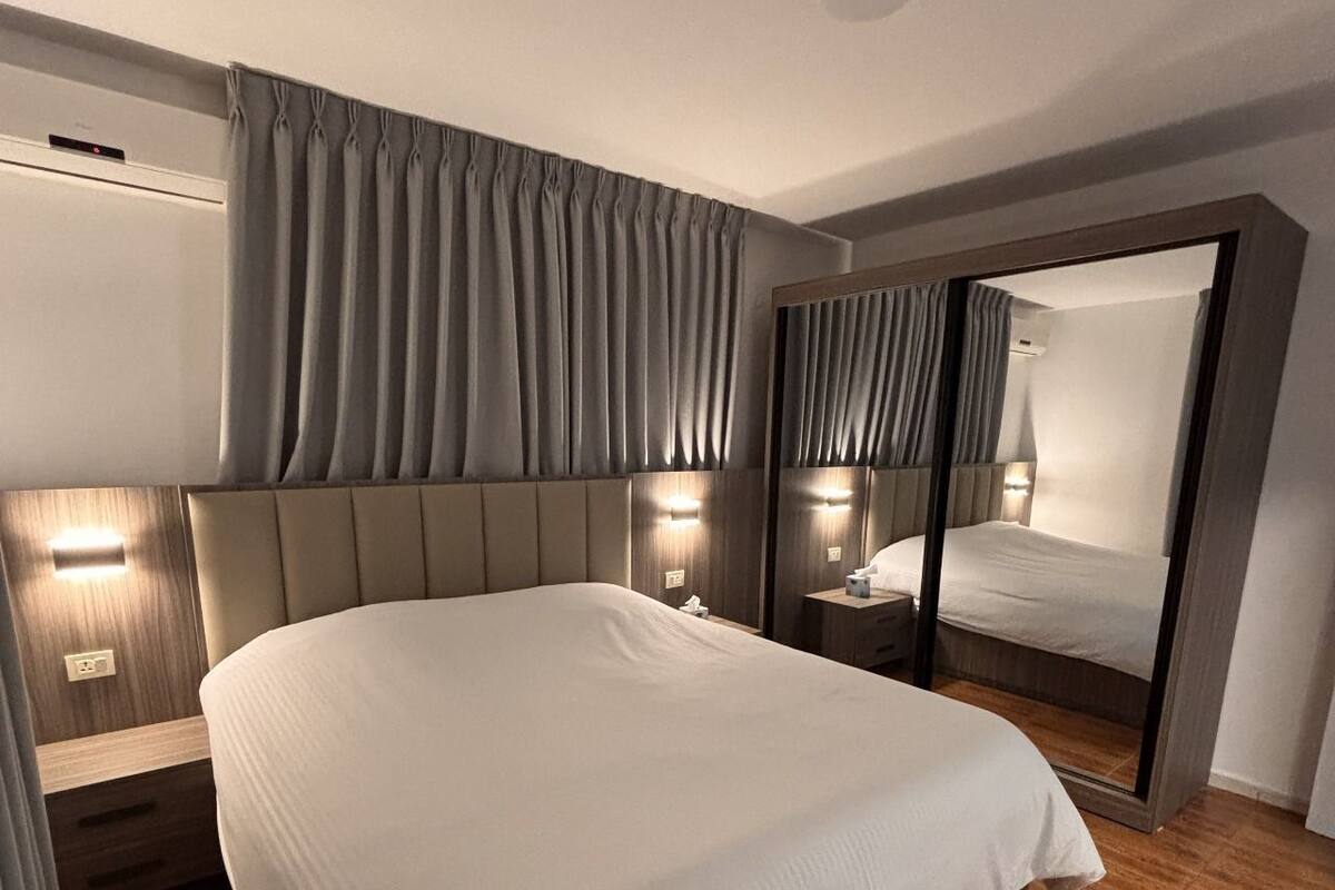 A modern hotel room with a large bed, a bedside table, a mirror, and a wardrobe.