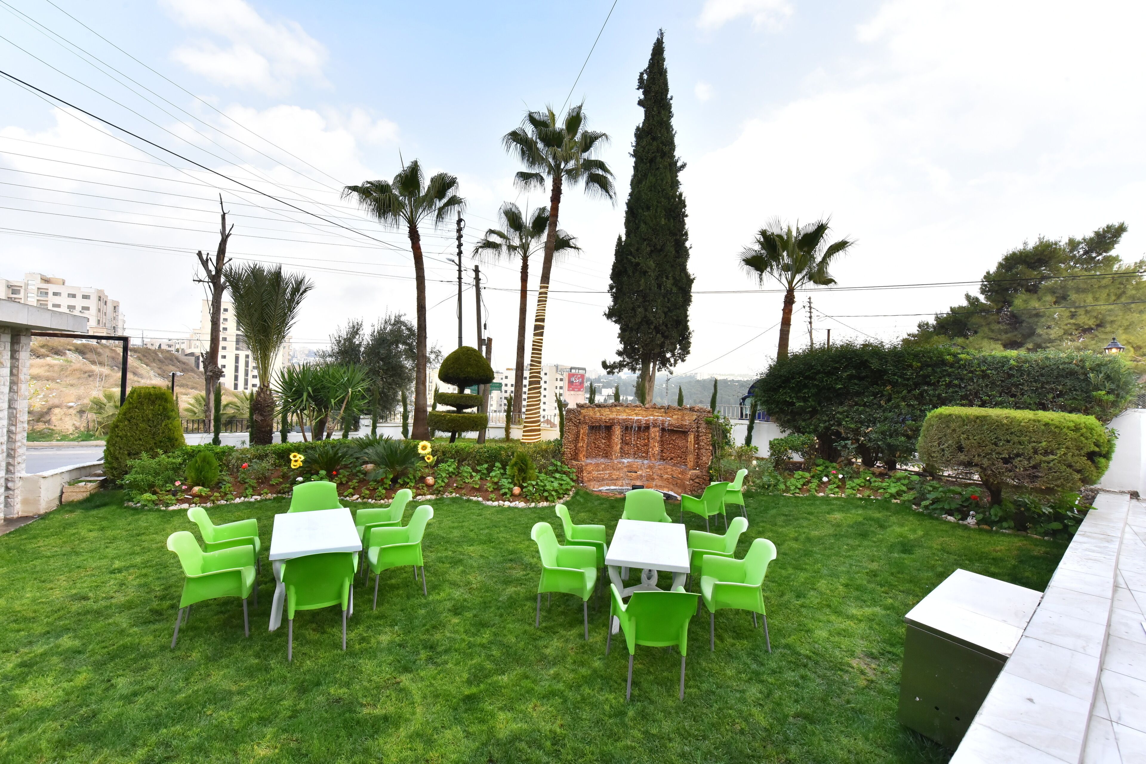 A well-maintained outdoor seating area with green chairs and white tables, surrounded by lush greenery and palm trees.