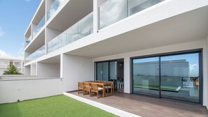 Un bâtiment moderne doté d’un toit plat, d’un balcon avec des garde-corps en verre et d’un petit espace pelousé agrémenté d’une table et de chaises en bois.