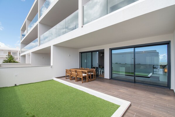 A modern building with a flat roof, a balcony with glass railings, and a small lawn area with a wooden table and chairs.