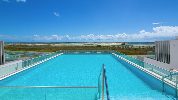 Une piscine d’un bleu éclatant, bordée d’une rambarde en verre, qui offre une vue sur une plage et l’océan.