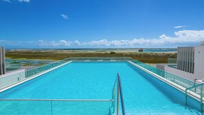 A clear blue pool with a glass railing, overlooking a beach and ocean.