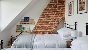 A bedroom with a sloped ceiling, a brick wall, a bed with white bedding, a bedside table with a lamp, and books on a shelf.